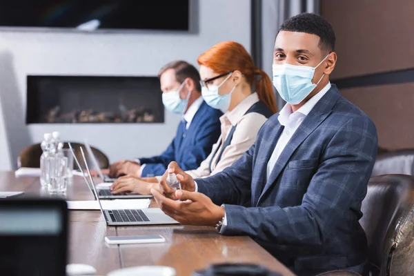 Hombre de negocios afroamericano en máscara médica mirando a la cámara y aplicando desinfectante mientras está sentado cerca de colegas en primer plano borroso - foto de stock