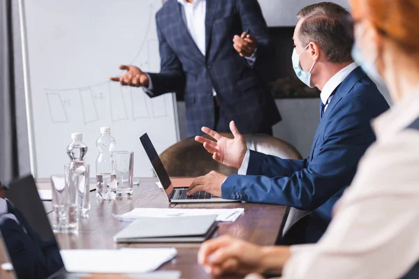 Businessman in medical mask gesturing near multicultural colleagues at workplace with digital devices on blurred foreground — Stock Photo