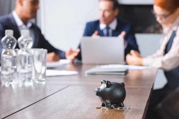 Small black piggy bank with coins on table with blurred multicultural businesspeople on background — Stock Photo
