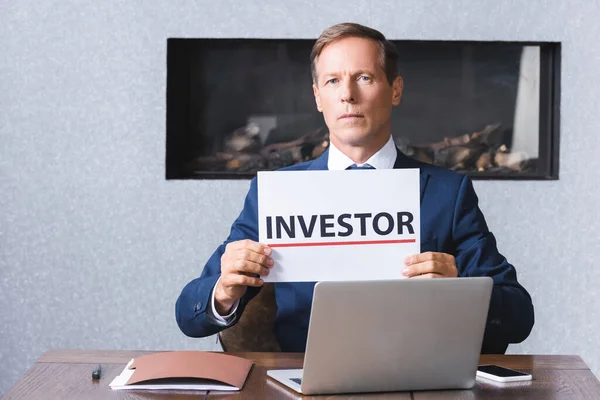 Homme d'affaires sérieux regardant la caméra et tenant signe avec le lettrage des investisseurs sur le lieu de travail avec des appareils numériques dans la salle de réunion — Photo de stock