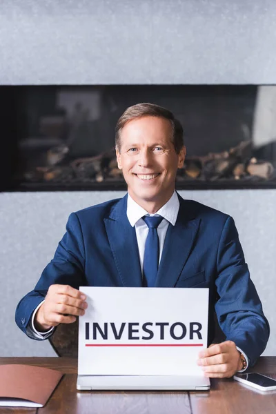 Homme d'affaires souriant regardant la caméra et tenant signe avec le lettrage de l'investisseur tout en étant assis sur le lieu de travail dans la salle de réunion — Photo de stock