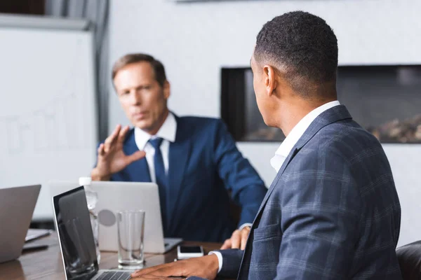 Uomo d'affari afroamericano seduto vicino esecutivo gesticolare sul posto di lavoro durante la riunione d'affari su sfondo sfocato — Foto stock