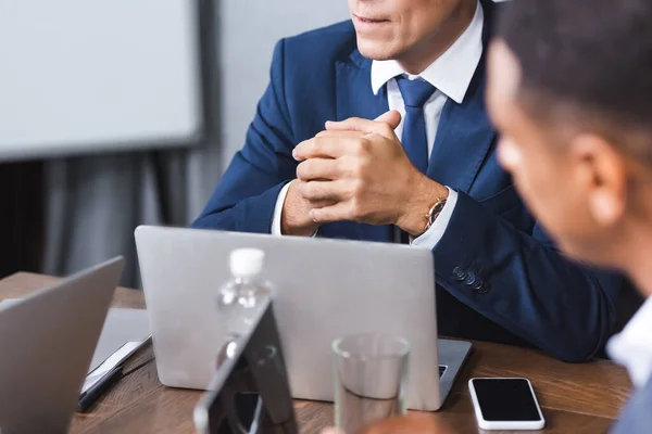 Executive with clenched hands sitting at workplace with digital devices near blurred african american businessman on foreground — Stock Photo