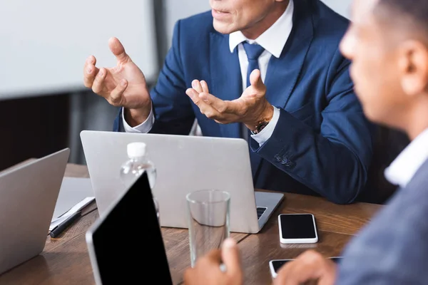 Vista recortada del gesto ejecutivo mientras está sentado en el lugar de trabajo cerca del borroso hombre de negocios afroamericano en primer plano - foto de stock