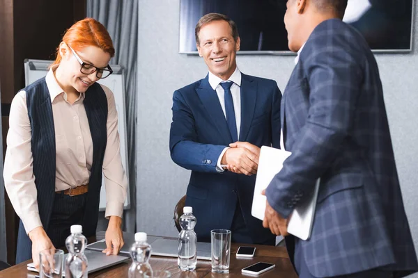 Smiling businessman shaking hands with african american colleague while standing near businesswoman in meeting room — Stock Photo