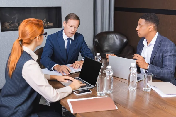 Executive seduta sul posto di lavoro con documenti e dispositivi digitali presso imprenditori multiculturali in sala riunioni — Foto stock