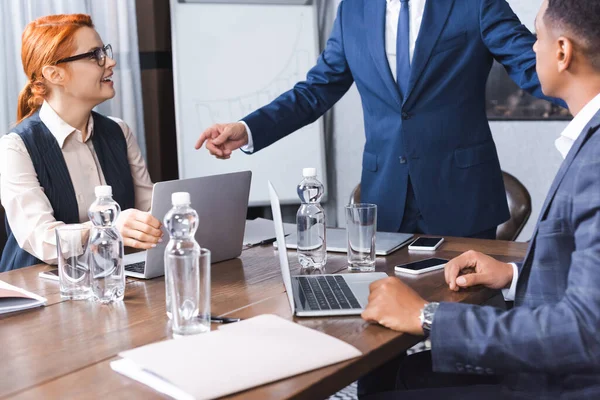 Colegas multiculturais sentados no local de trabalho com dispositivos digitais perto de empresário de pé e gesticulando na sala de reuniões — Fotografia de Stock