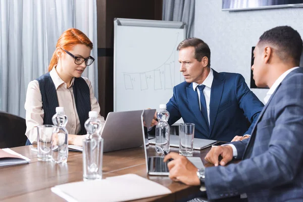 Multicultural businessmen looking at laptop near redhead businesswoman at workplace in boardroom — Stock Photo