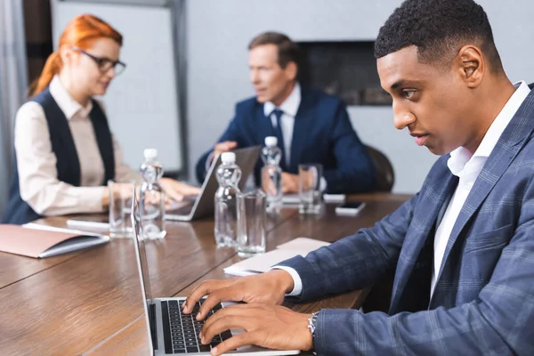 Concentrated african american businessman typing on laptop at workplace with blurred colleagues on background — Stock Photo