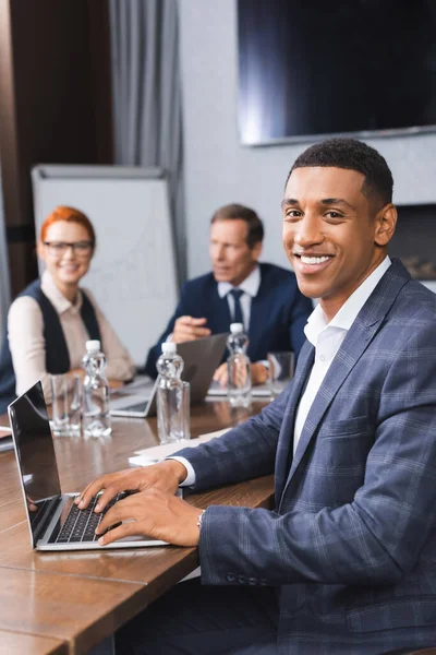 Sorrindo empresário afro-americano olhando para a câmera enquanto digita no laptop com colegas embaçados no fundo — Fotografia de Stock