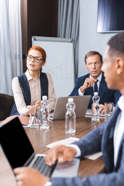 Redhead businesswoman talking while sitting at workplace with multicultural colleagues on blurred foreground in meeting room — Stock Photo