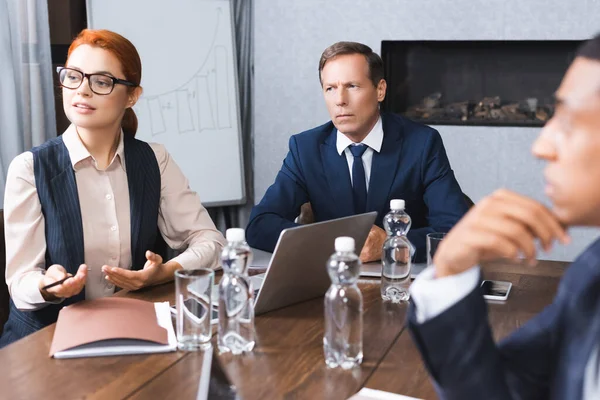 Femme d'affaires rousse gesticulant tout en étant assis sur le lieu de travail avec un collègue afro-américain flou au premier plan — Photo de stock