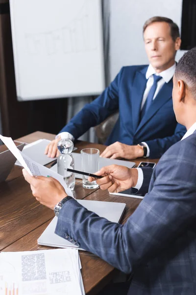 Empresario afroamericano apuntando con bolígrafo al documento mientras está sentado cerca de un ejecutivo reflexivo sobre fondo borroso - foto de stock