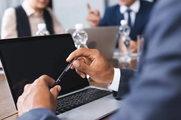 Vista recortada del empresario afroamericano sosteniendo la pluma cerca de la computadora portátil con colegas borrosos en el fondo - foto de stock