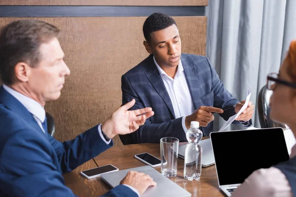 Uomo d'affari afroamericano che punta il dito contro la carta mentre siede sul posto di lavoro vicino a colleghi sfocati in primo piano — Foto stock