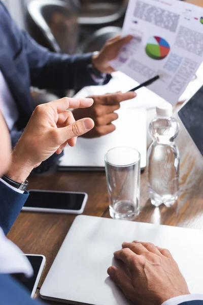 Vista recortada del hombre de negocios señalando con el dedo en el papel con gráfico con colega borroso en el fondo — Stock Photo