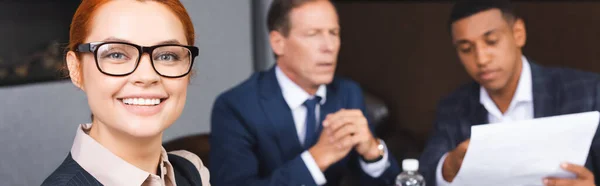 Happy redhead businesswoman looking at camera with blurred multicultural colleagues on background, banner — Stock Photo