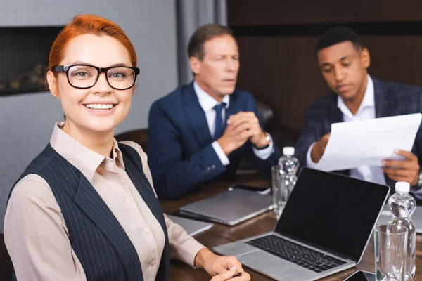 Smiling businesswoman looking at camera while sitting at workplace with digital devices and blurred colleagues on background — Stock Photo