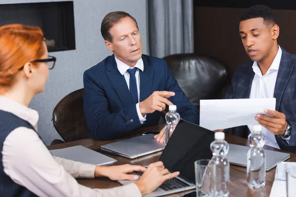 Ejecutivo señalando con el dedo en el papel en manos de empresario afroamericano en el lugar de trabajo en primer plano borroso - foto de stock