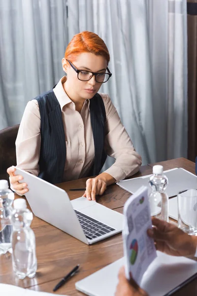 Redhead businesswoman holding laptop while sitting at workplace with blurred colleague on foreground in meeting room — Stock Photo