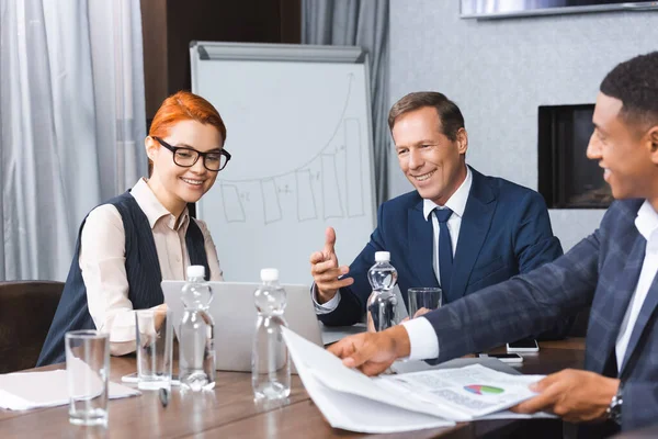 Smiling businesspeople looking at laptop while sitting at workplace near African american businessman with documents - foto de stock