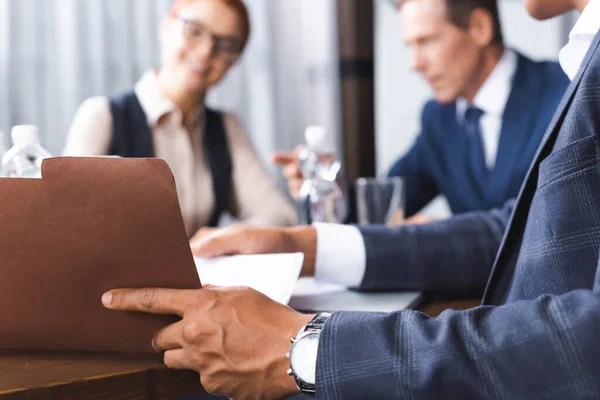 Close up view of african american businessman opening paper folder at workplace with blurred businesspeople on background — Stock Photo