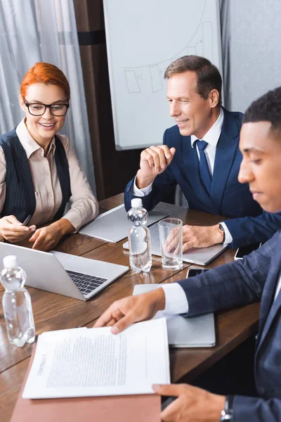 Donna d'affari sorridente che punta con penna al computer portatile vicino investitore sul posto di lavoro con l'uomo africano americano offuscato in primo piano — Foto stock