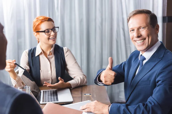 Smiling executive with thumb up looking at camera at workplace with laptop near businesspeople on blurred foreground — Stock Photo