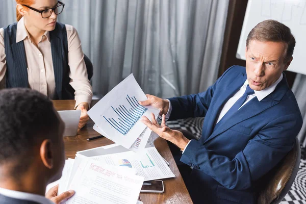 Ansicht eines irritierten Managers mit Papieren, die gestikulieren, während er den afrikanisch-amerikanischen Geschäftsmann im verschwommenen Vordergrund betrachtet — Stockfoto