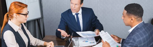 Dissatisfied executive with documents sitting near multicultural colleagues at workplace in meeting room, banner — Stock Photo