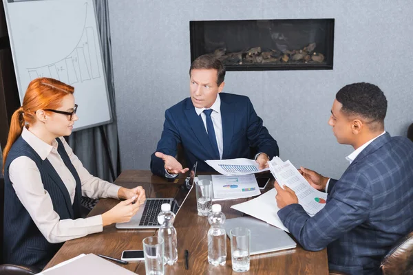 Ejecutivo irritado con documentos que señalan mientras mira a la mujer de negocios sentada en el lugar de trabajo cerca de un colega afroamericano - foto de stock