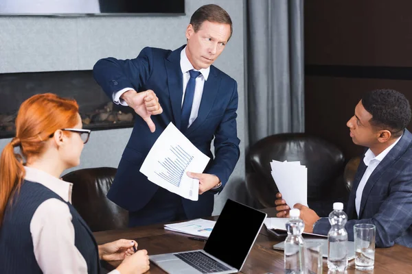 Dissatisfied executive with papers showing thumb down while looking at african american businessman at workplace — Stock Photo