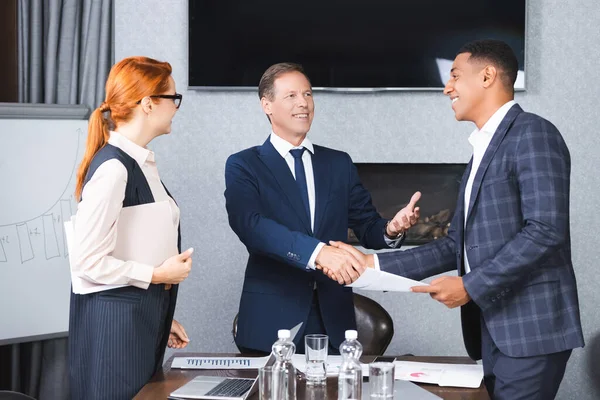 Felices hombres de negocios multiculturales dándose la mano mientras están cerca de la pelirroja empresaria en la sala de juntas - foto de stock