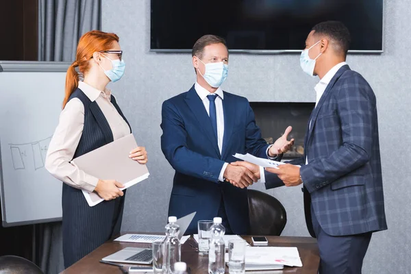 Мультикультурные бизнесмены в медицинских масках пожимают друг другу руки, стоя рядом с предпринимательницей в зале заседаний — стоковое фото