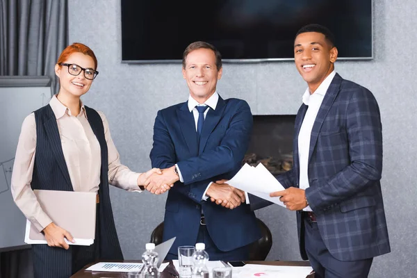 Happy businessman looking at camera while shaking hands with multicultural colleagues near workplace in boardroom — Stock Photo