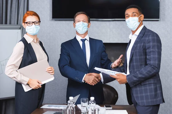 Багатокультурні бізнесмени в медичних масках трясуться один з одним, стоячи поруч з бізнес-леді в залі засідань — стокове фото