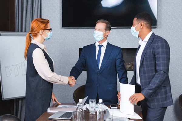 Geschäftsleute in medizinischen Masken schütteln einander die Hände, während sie neben einem Afroamerikaner im Sitzungssaal stehen — Stockfoto