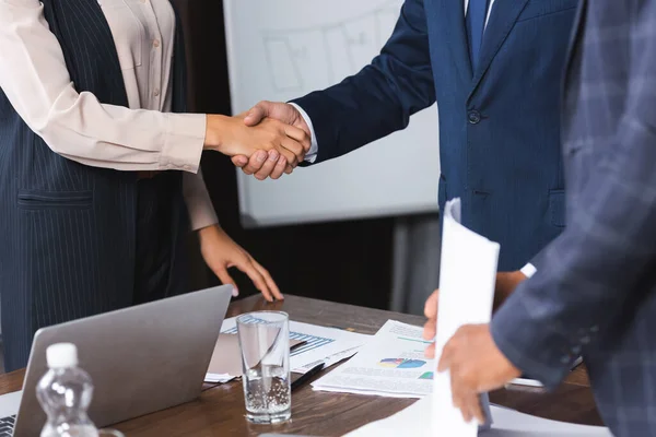 Cropped view of businesspeople shaking hands with each other near blurred african american man on foreground in boardroom — Stock Photo