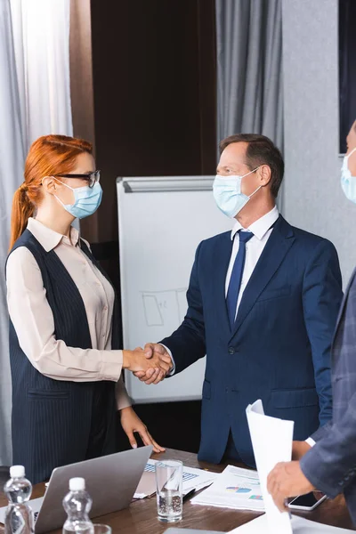 Businesspeople in medical masks shaking hands with each other near blurred african american man on foreground in boardroom — Stock Photo