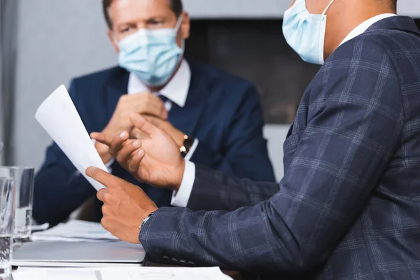 Африканський американський бізнесмен у медичній масці вказує пальцем на папір на робочому місці з розмитим колегою на задньому плані. — стокове фото