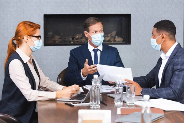 Multicultural businesspeople in medical masks talking while sitting at workplace with devices and papers on blurred foreground — Stock Photo