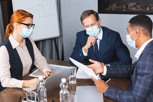 Multicultural businesspeople in medical masks talking while sitting at workplace with digital devices and documents — Stock Photo