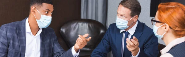 Multicultural businesspeople in medical masks gesturing during discussion near female colleague in boardroom, banner — Stock Photo