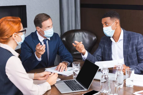 Multicultural businesspeople in medical masks gesturing during discussion near female colleague at workplace, banner — Stock Photo