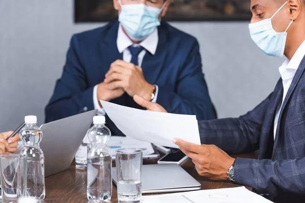 African american businessman in medical mask showing papers while sitting at workplace with blurred colleague on background — Stock Photo