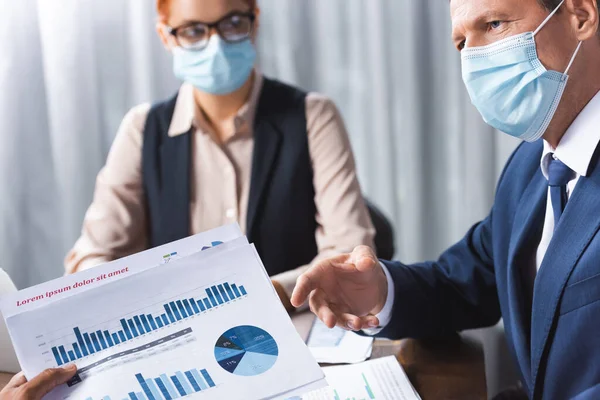 Бизнесмен в медицинской маске жестом и глядя на коллегу показывает документы с графиками на размытом фоне — стоковое фото