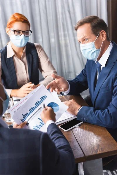 Businessman in medical mask looking at african american colleague showing papers with charts on blurred foreground — Stock Photo