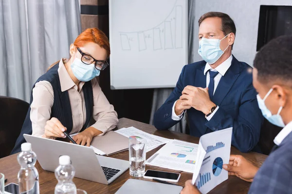 Donna d'affari in maschera medica che punta con penna al computer portatile vicino investitore con collega afroamericano offuscata in primo piano — Foto stock