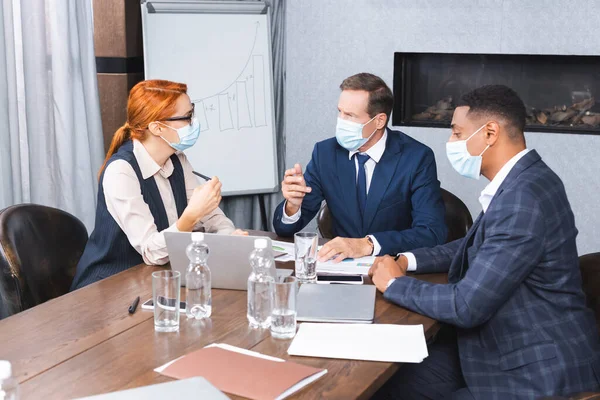 Empresarios multiculturales con máscaras médicas hablando con inversionistas mientras están sentados en el lugar de trabajo con dispositivos digitales - foto de stock