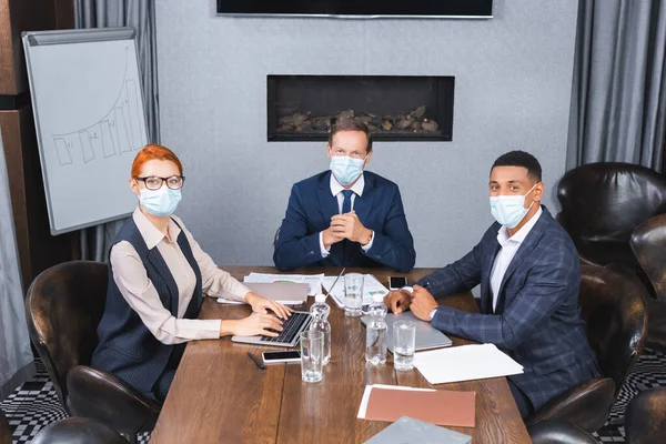 Empresarios multiculturales con máscaras médicas mirando a la cámara mientras están sentados en el lugar de trabajo con dispositivos digitales en la sala de reuniones - foto de stock